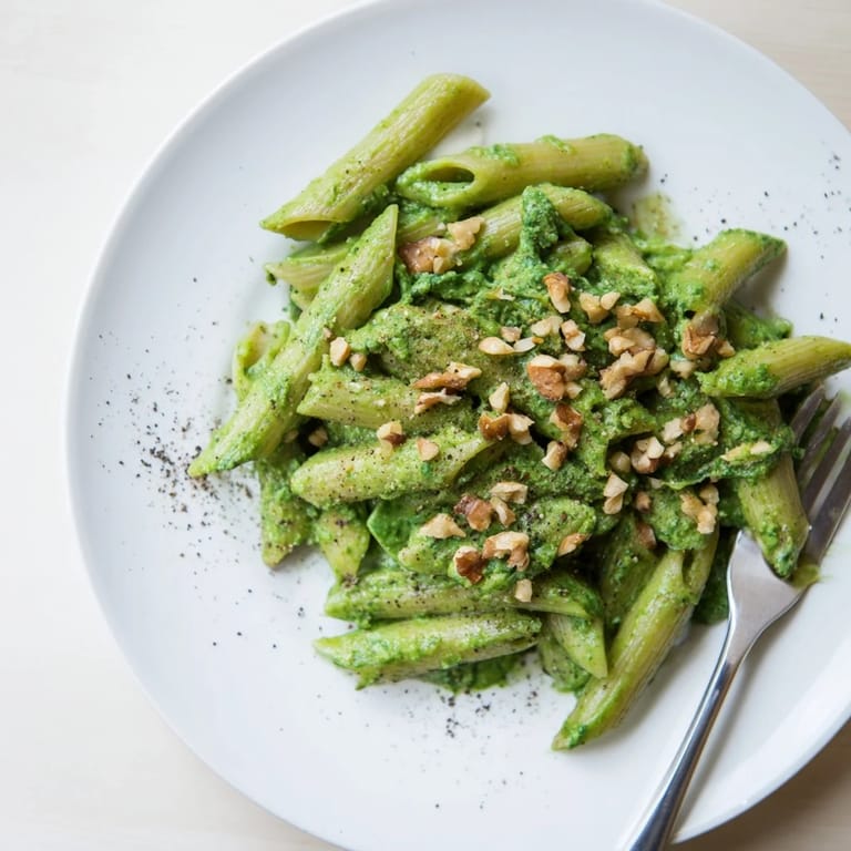 A white ceramic bowl of creamy spinach walnut pasta, steam rising from the velvety green sauce, with fresh spinach leaves and chopped walnuts nearby.